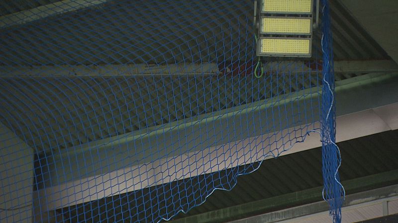Red de seguridad azul instalada bajo las vigas de hormigón y la cubierta del techo de un estadio, junto a un foco de iluminación LED.