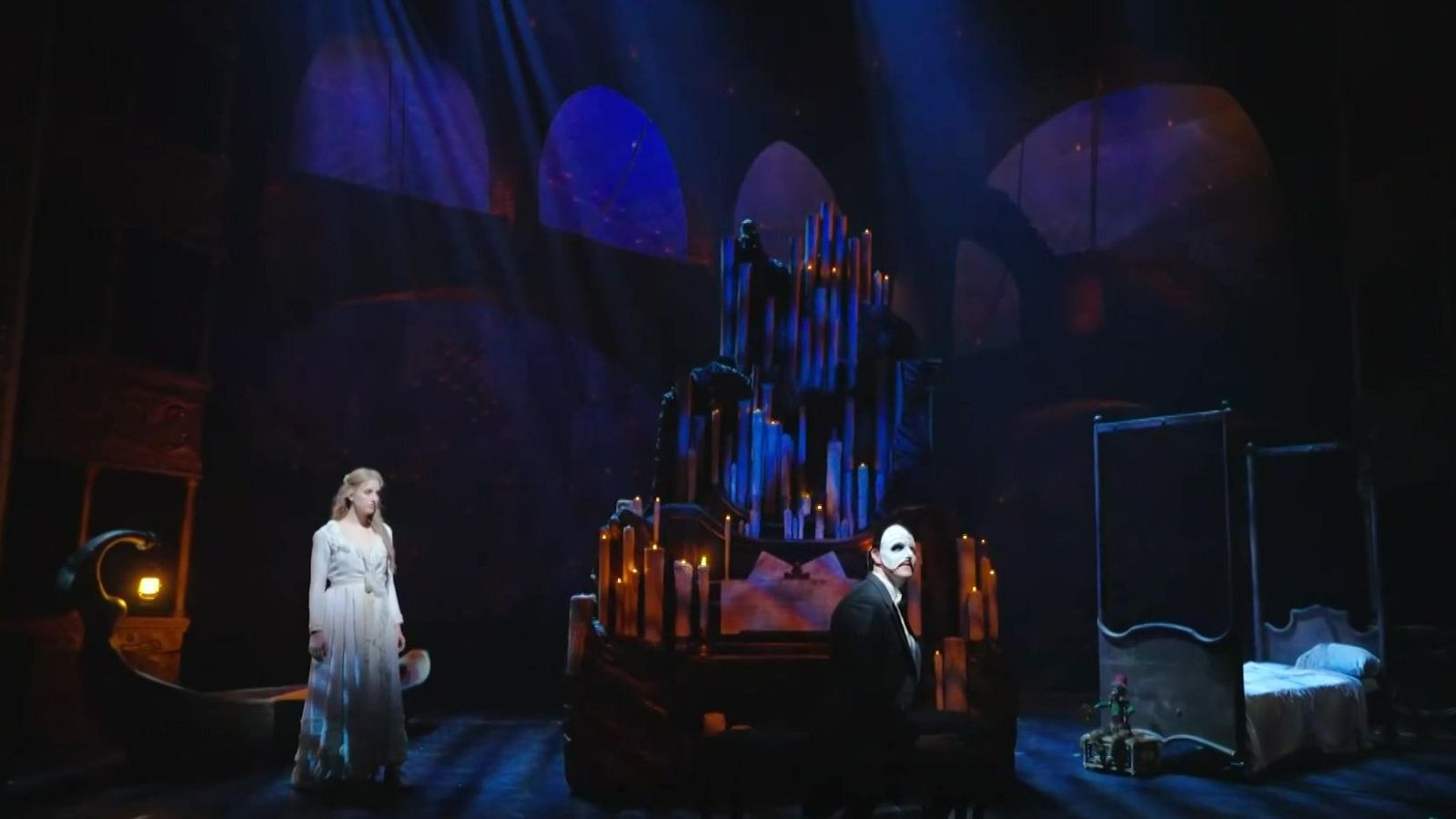 Escena del musical "El Fantasma de la Ópera" con Christine Daaé y el Fantasma, con su icónica máscara, junto a un órgano iluminado por velas en una atmósfera gótica.