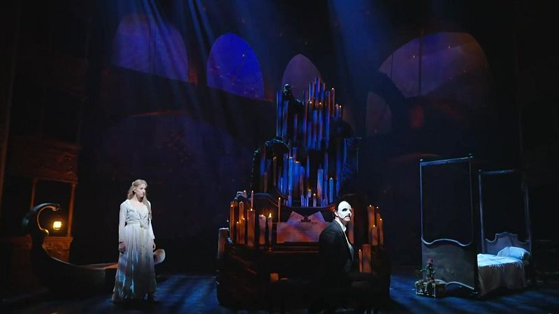Escena del musical "El Fantasma de la Ópera" con Christine Daaé y el Fantasma, con su icónica máscara, junto a un órgano iluminado por velas en una atmósfera gótica.