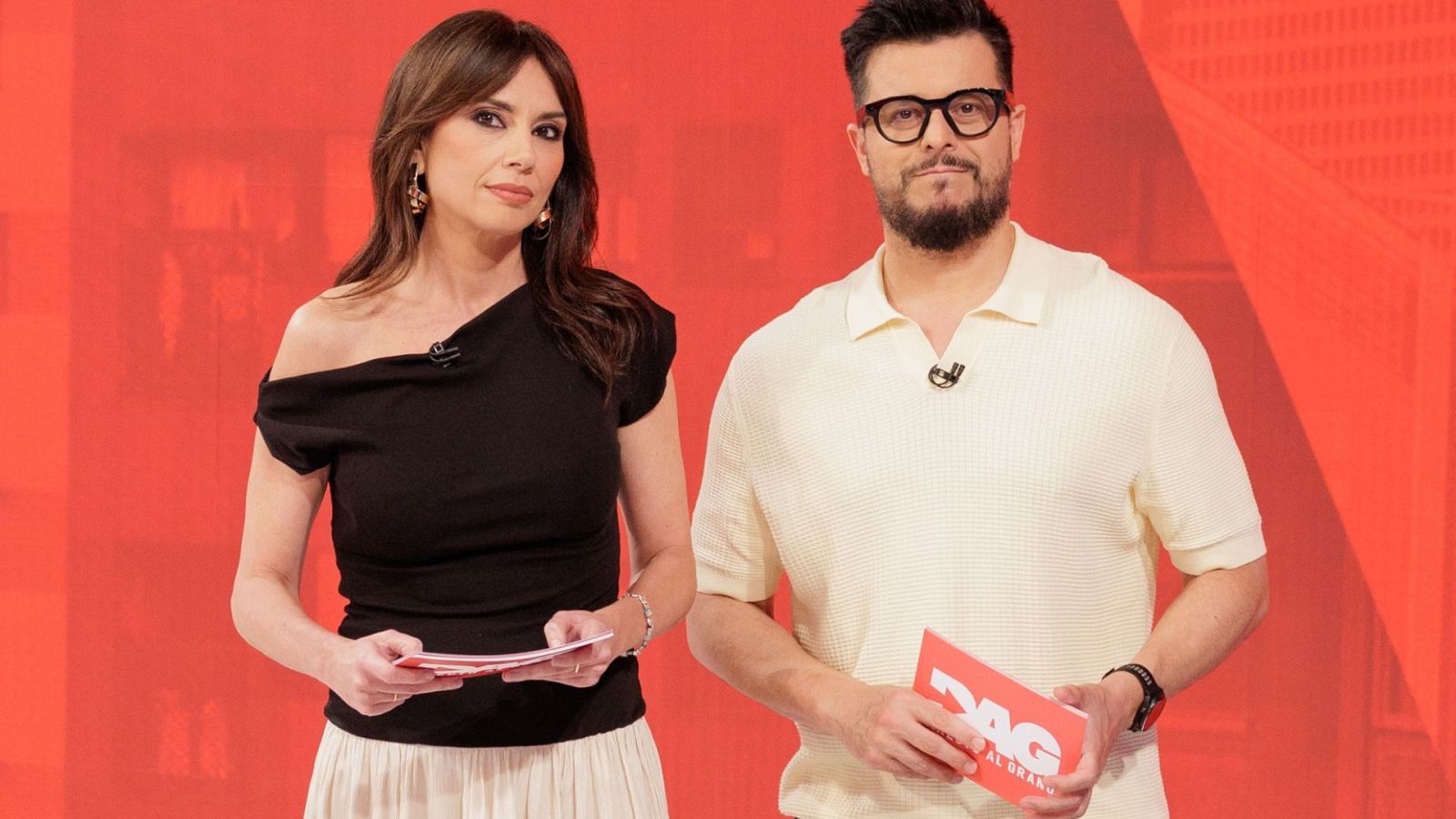 Los presentadores Alba Lancha y Toni Díaz posan en el plató de "Directo al grano", sosteniendo tarjetas del programa con las siglas "DAG" sobre un fondo rojo con formas geométricas.