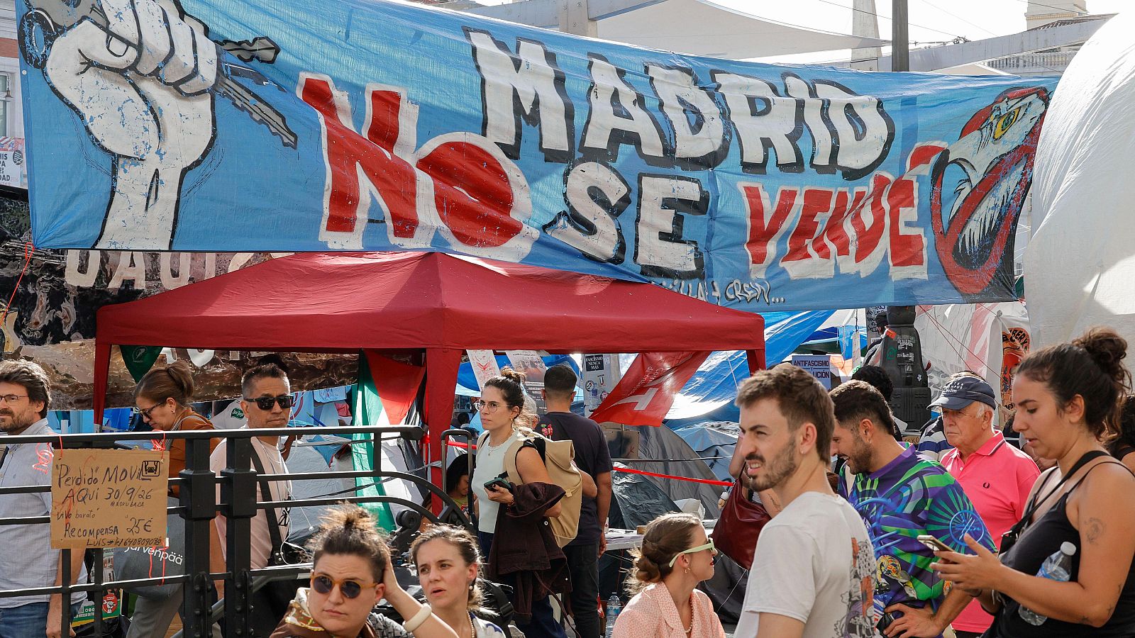 Protesta ciudadana bajo una gran pancarta que reza "MADRID NO SE VENDE", con personas reunidas junto a una carpa roja y un cartel manuscrito que ofrece recompensa por un móvil perdido.
