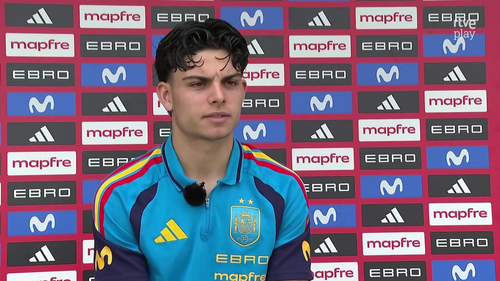 El futbolista Juanlu Sánchez atiende a los medios con la ropa de la selección, luciendo un polo azul celeste con los escudos de la RFEF y marcas patrocinadoras.