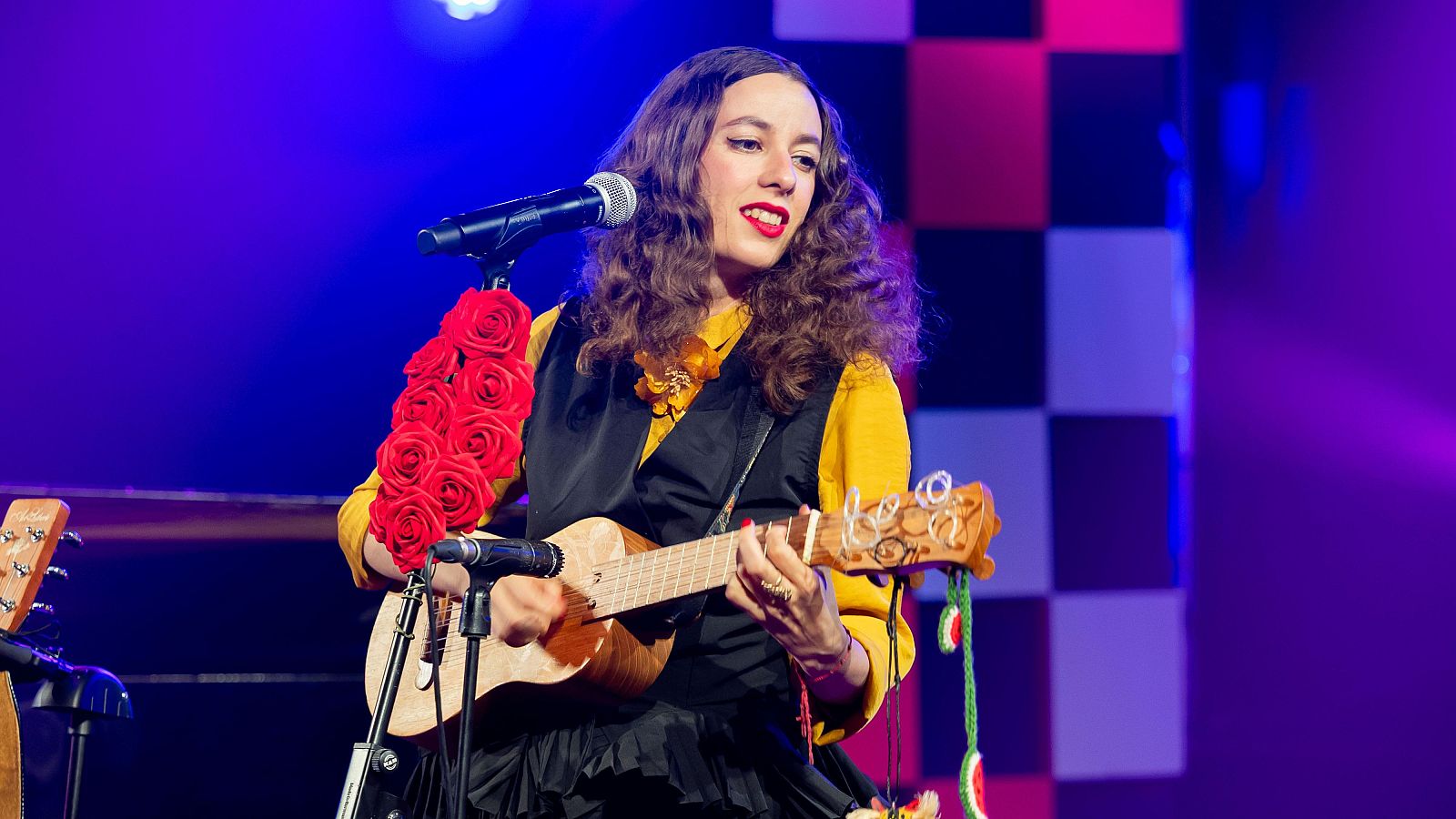 Sofía Comas canta ante un micrófono decorado con rosas rojas mientras toca un pequeño instrumento acústico con detalles de ganchillo.