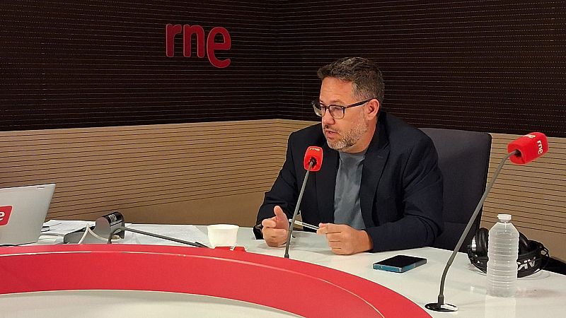 Hombre con barba y gafas sentado en la mesa de un estudio de radio, rodeado de micrófonos con espuma roja, un móvil y una botella de agua.