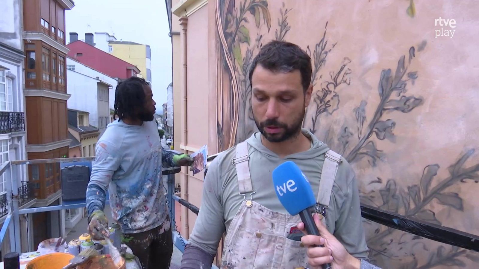 Dos artistas sobre una plataforma elevadora crean un mural urbano mientras uno es entrevistado con un micrófono de tve.