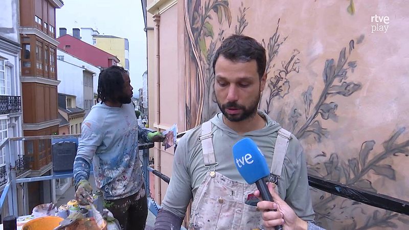 Dos artistas sobre una plataforma elevadora crean un mural urbano mientras uno es entrevistado con un micrófono de tve.