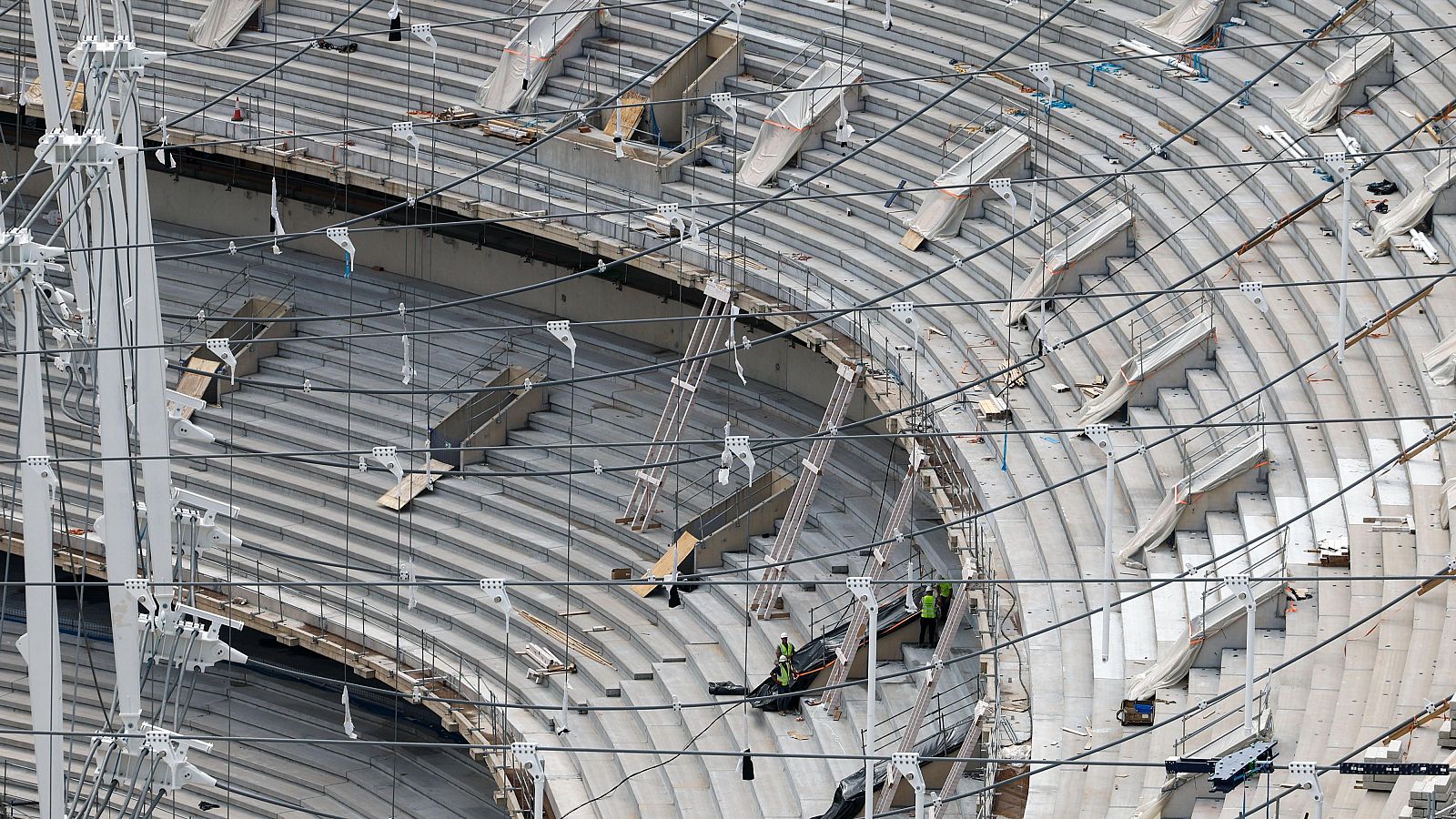 Graderío de hormigón de un estadio en construcción con cables de acero tensados para la cubierta y operarios trabajando.