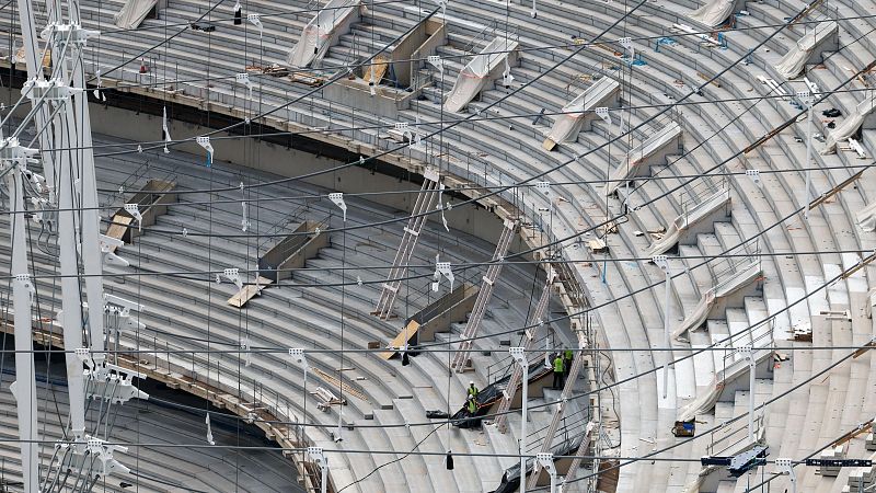 Graderío de hormigón de un estadio en construcción con cables de acero tensados para la cubierta y operarios trabajando.