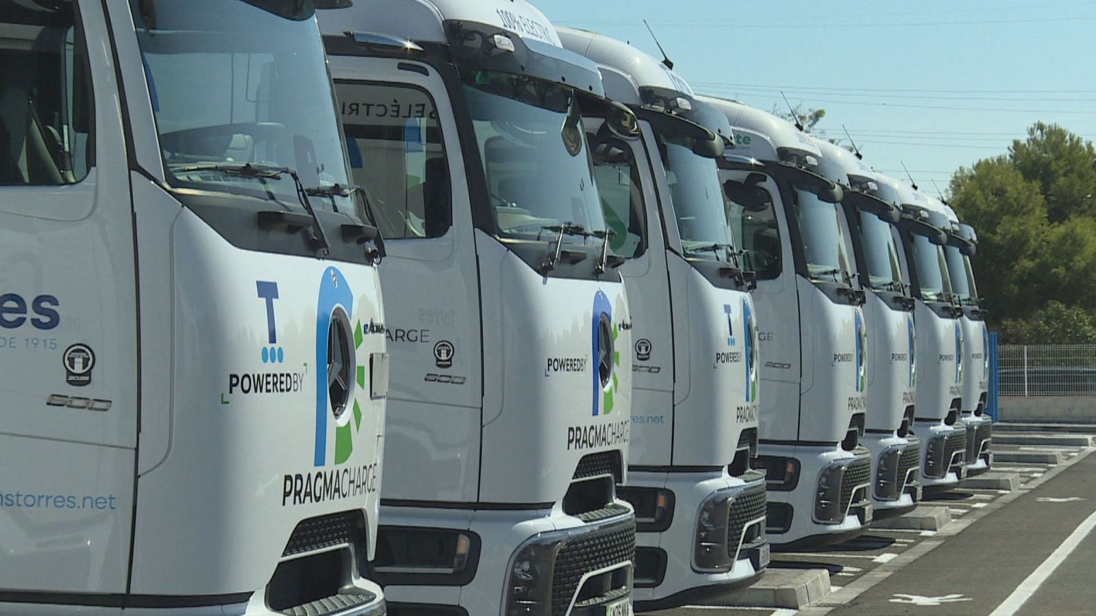 Hilera de cabezas tractoras de camiones blancos y eléctricos aparcadas en un área logística al aire libre con diseño aerodinámico y cámaras digitales.
