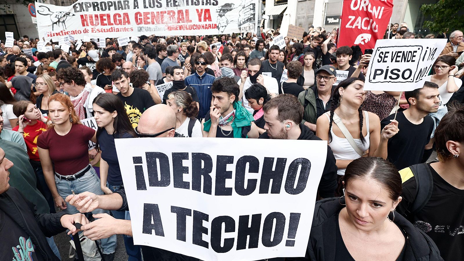 Manifestación urbana diurna de jóvenes con pancartas reivindicativas como «¡DERECHO A TECHO!» y «SE VENDE PISO ¡YA NO!».