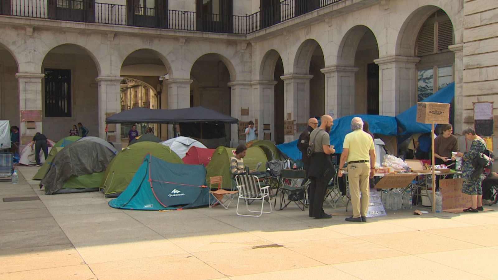 Campamento de protesta con tiendas, sillas plegables y una zona de intendencia con provisiones instalado en una plaza porticada pavimentada.