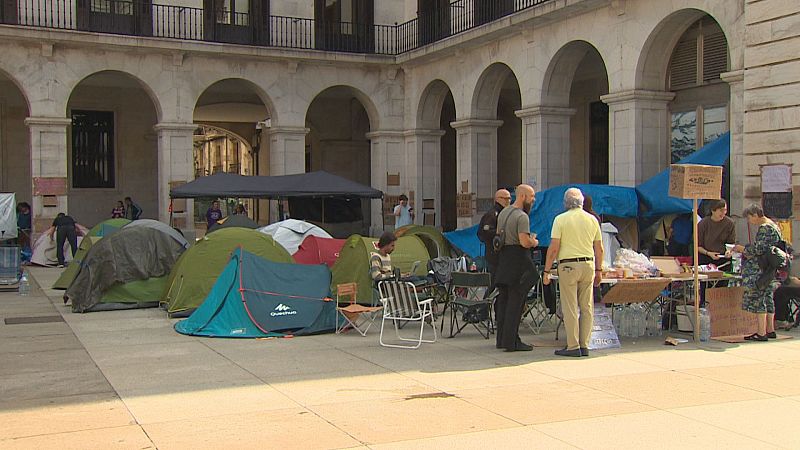 Campamento de protesta con tiendas, sillas plegables y una zona de intendencia con provisiones instalado en una plaza porticada pavimentada.