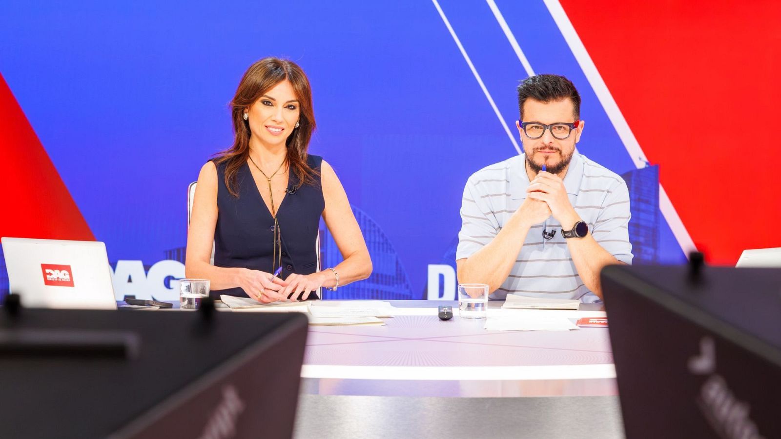 Dos presentadores de televisión están sentados ante una mesa en un plató con paneles azules y rojos, sosteniendo bolígrafos junto a papeles y vasos de agua.