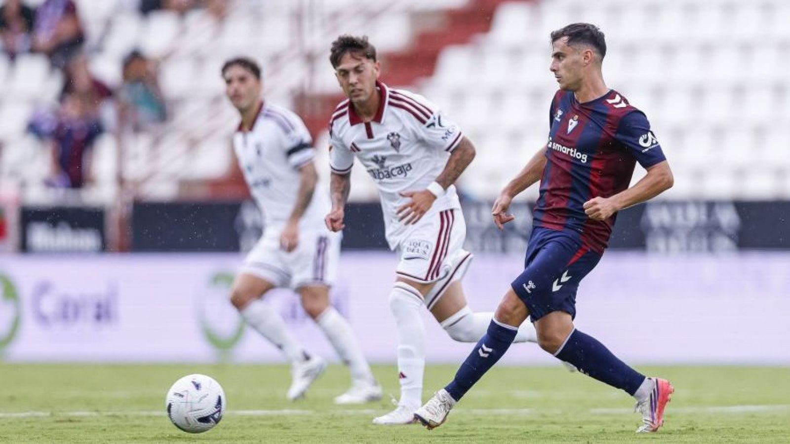 Futbolista con equipación azulgrana de la SD Eibar corre con el balón mientras dos jugadores del Albacete Balompié le persiguen en el campo.