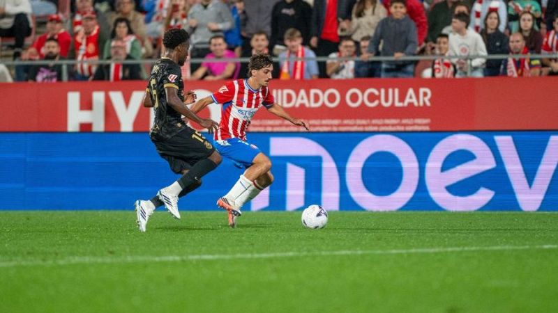 El futbolista Rodrigo Riquelme conduce el balón con la camiseta rojiblanca del Girona FC mientras un rival de negro le persigue.