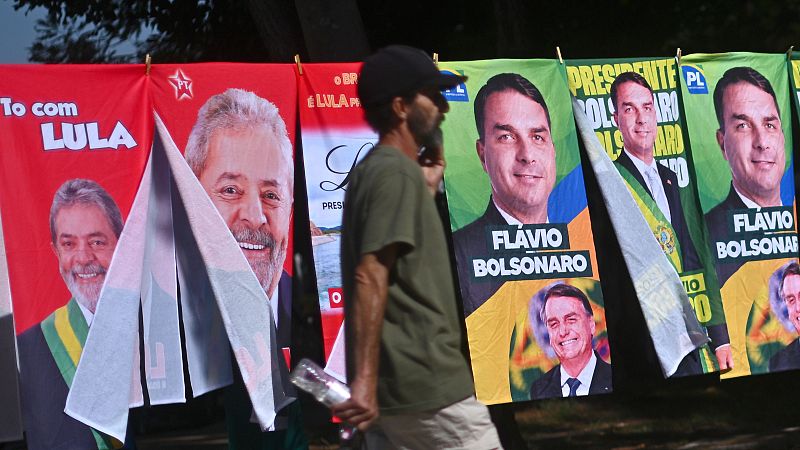 Hombre camina frente a un puesto callejero con toallas de campaña electoral de Lula da Silva y Bolsonaro.