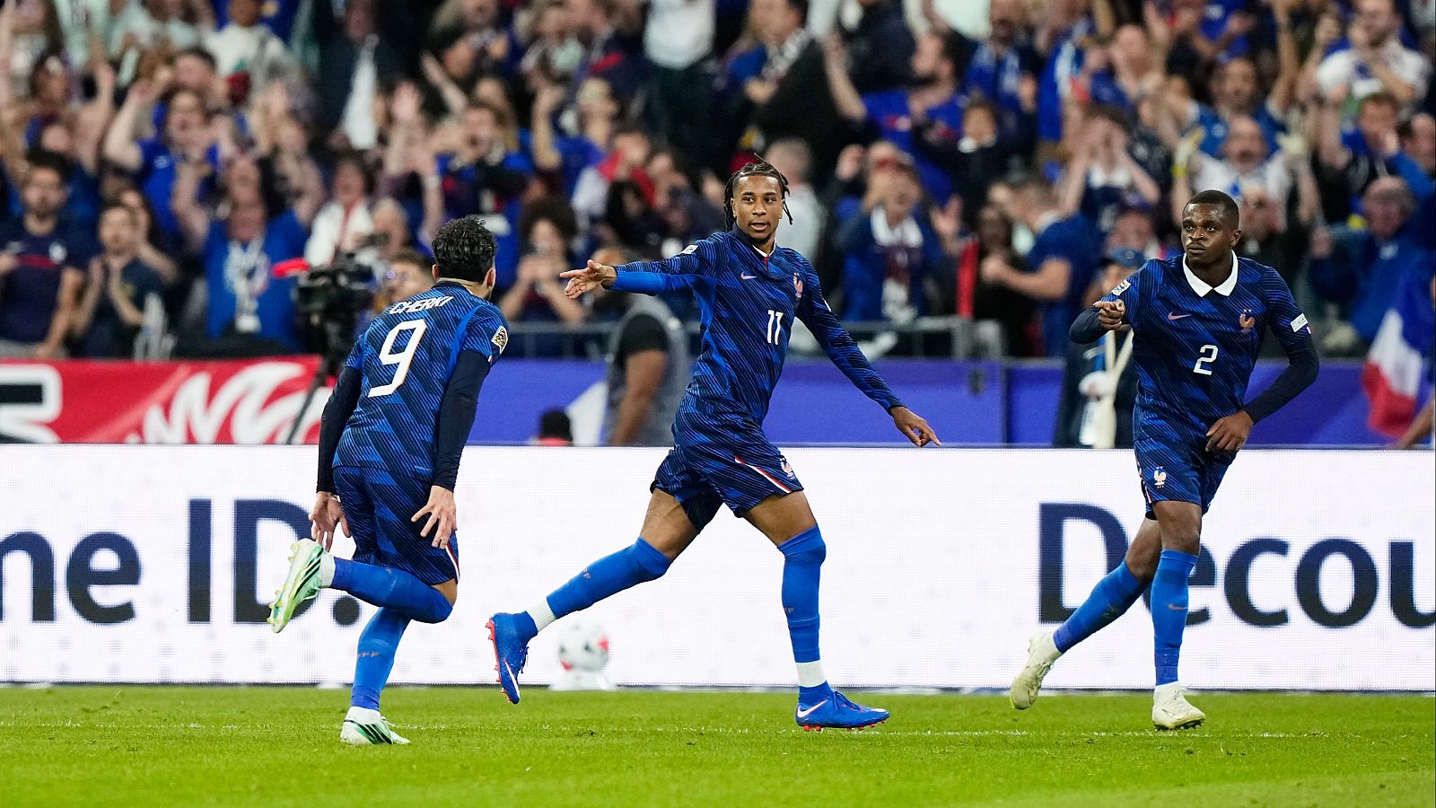 Tres futbolistas franceses celebran eufóricos sobre el césped verde de un estadio repleto de aficionados que ondean banderas y visten de azul.