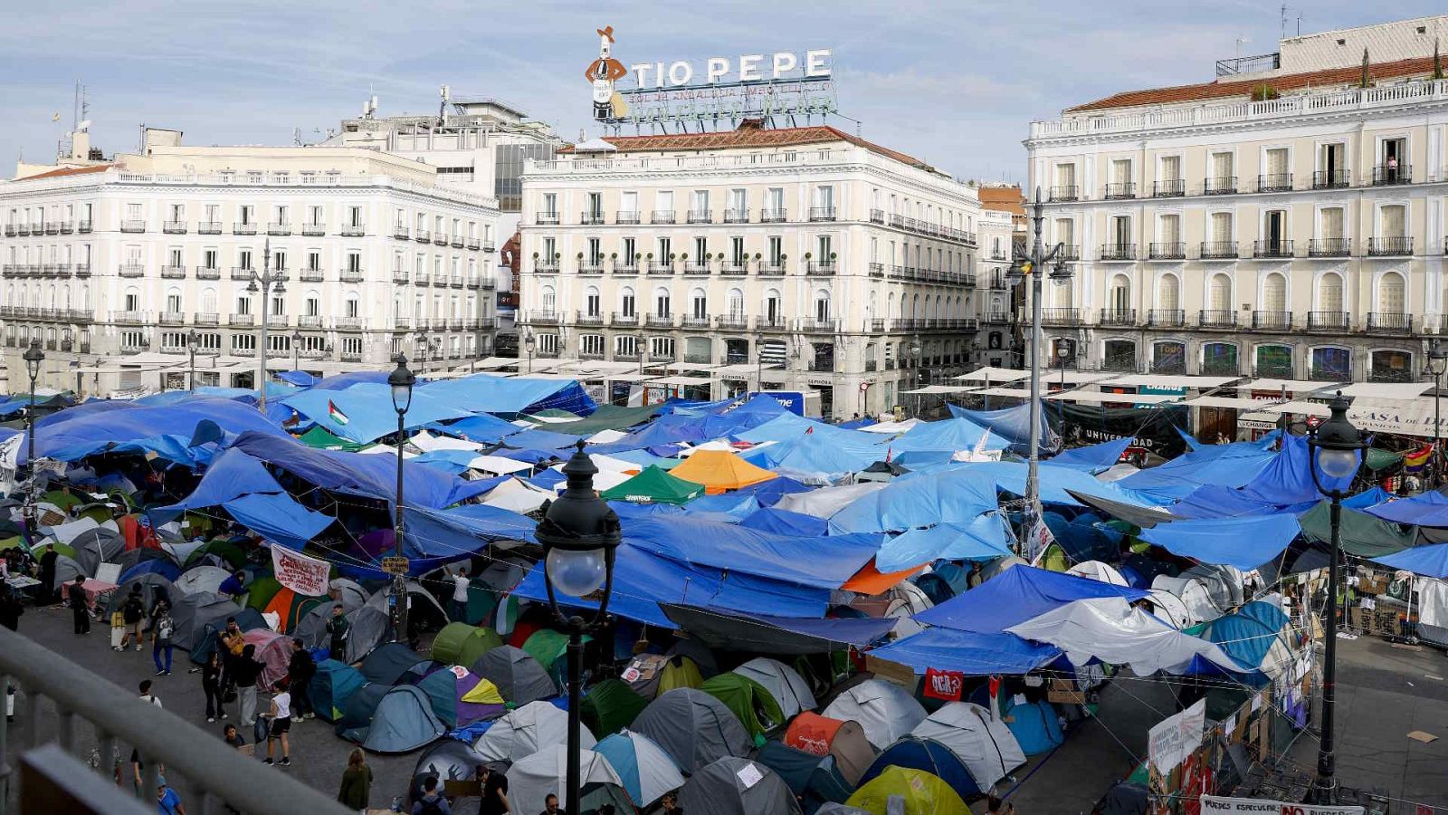 Vista aérea de la Puerta del Sol en Madrid con un gran campamento de protesta formado por tiendas y toldos, destacando el cartel de Tío Pepe al fondo.