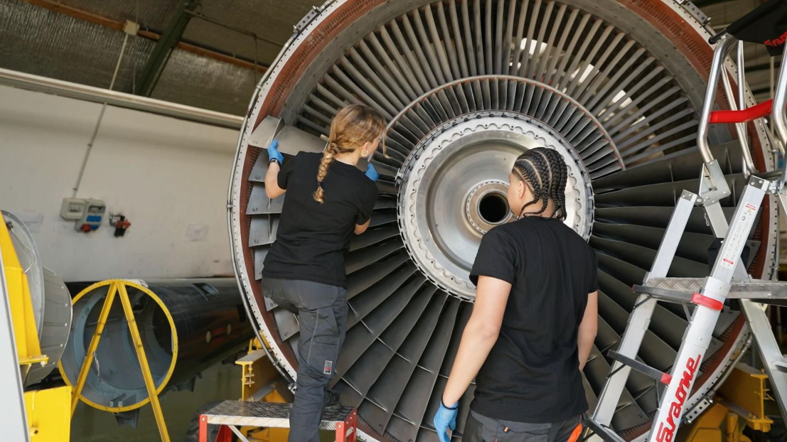 Dos jóvenes estudiantes revisan un gigantesco motor de avión en un taller industrial, inspeccionando los álabes metálicos de la turbina con guantes protectores.