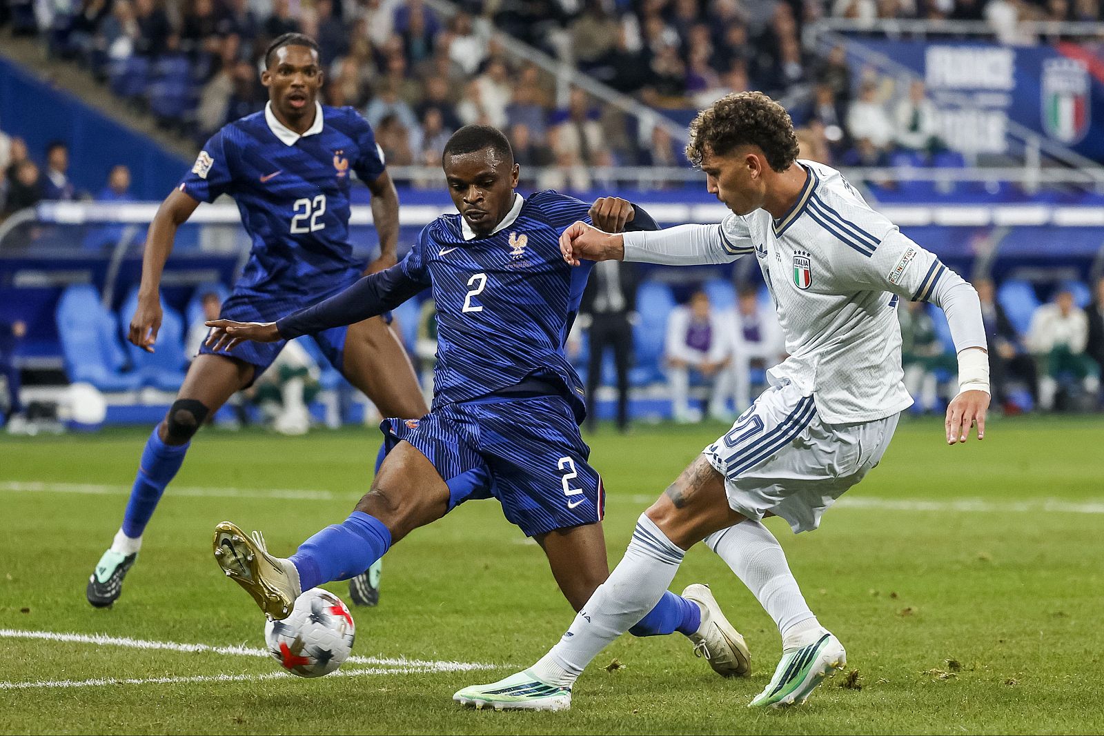 Intensa disputa de balón a ras de césped entre un jugador francés con la camiseta número 2 y un rival italiano durante un partido de fútbol.