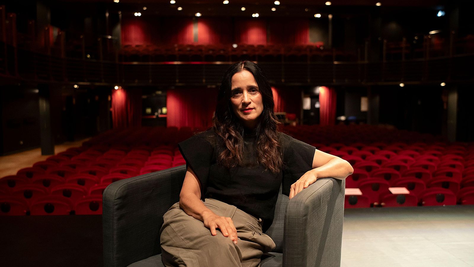 La cantautora Julieta Venegas está sentada en un sillón gris en el escenario de un teatro con butacas rojas.