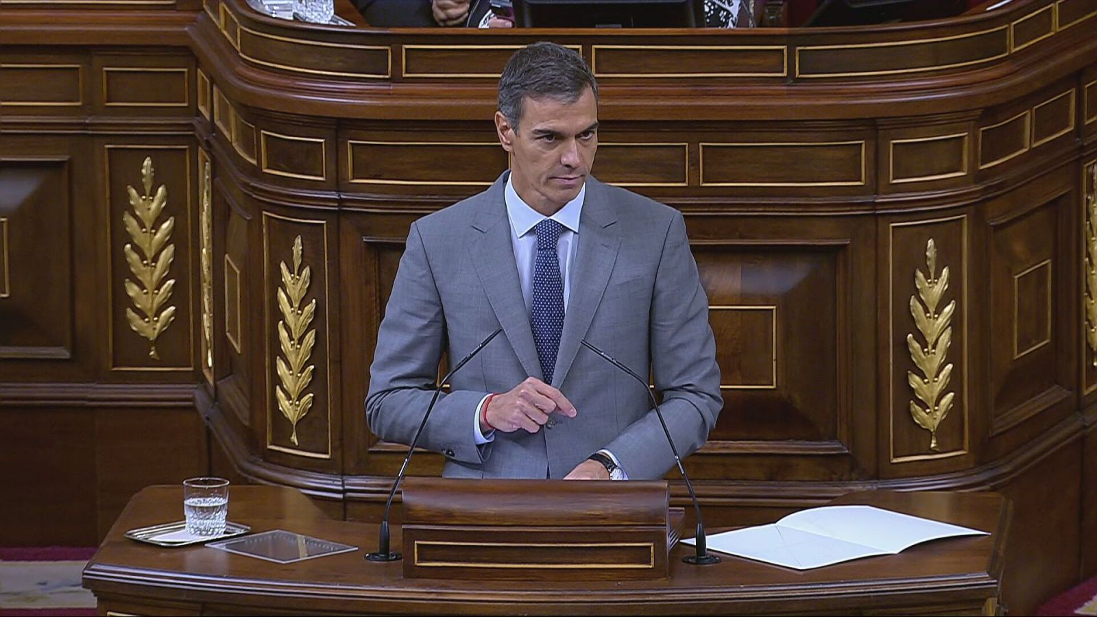 Plano medio de Pedro Sánchez interviniendo en la tribuna del Congreso, vestido con traje gris y corbata azul ante un atril de madera.