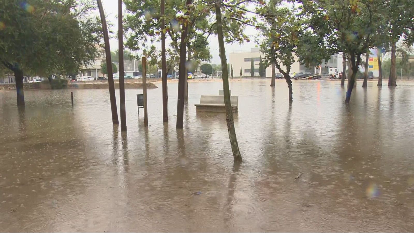 Parque urbano anegado por el agua tras fuertes lluvias, con un banco de piedra parcialmente sumergido y vehículos al fondo.