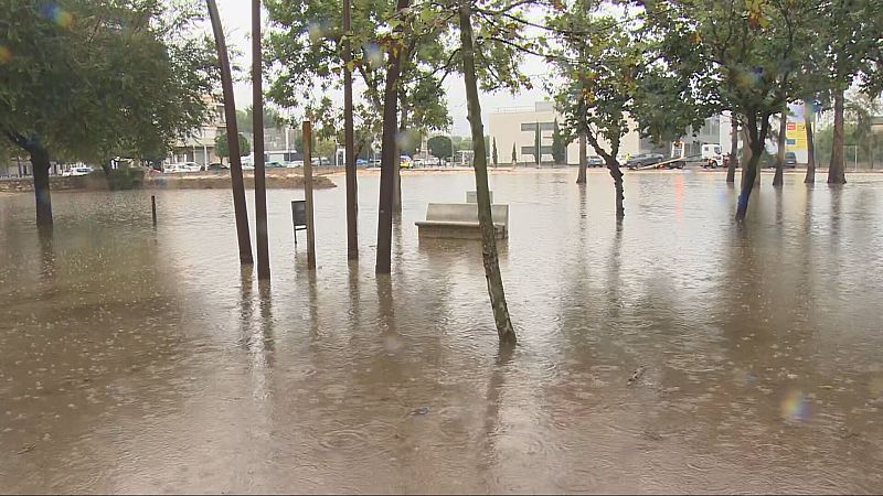Parque urbano anegado por el agua tras fuertes lluvias, con un banco de piedra parcialmente sumergido y vehículos al fondo.