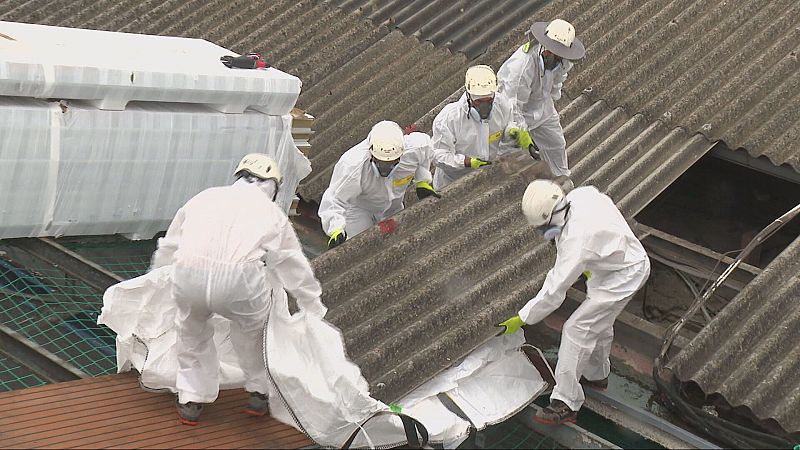 Cinco operarios con EPIs blancos trabajan en equipo sobre una cubierta inclinada retirando placas de fibrocemento y utilizando una saca industrial para los residuos.