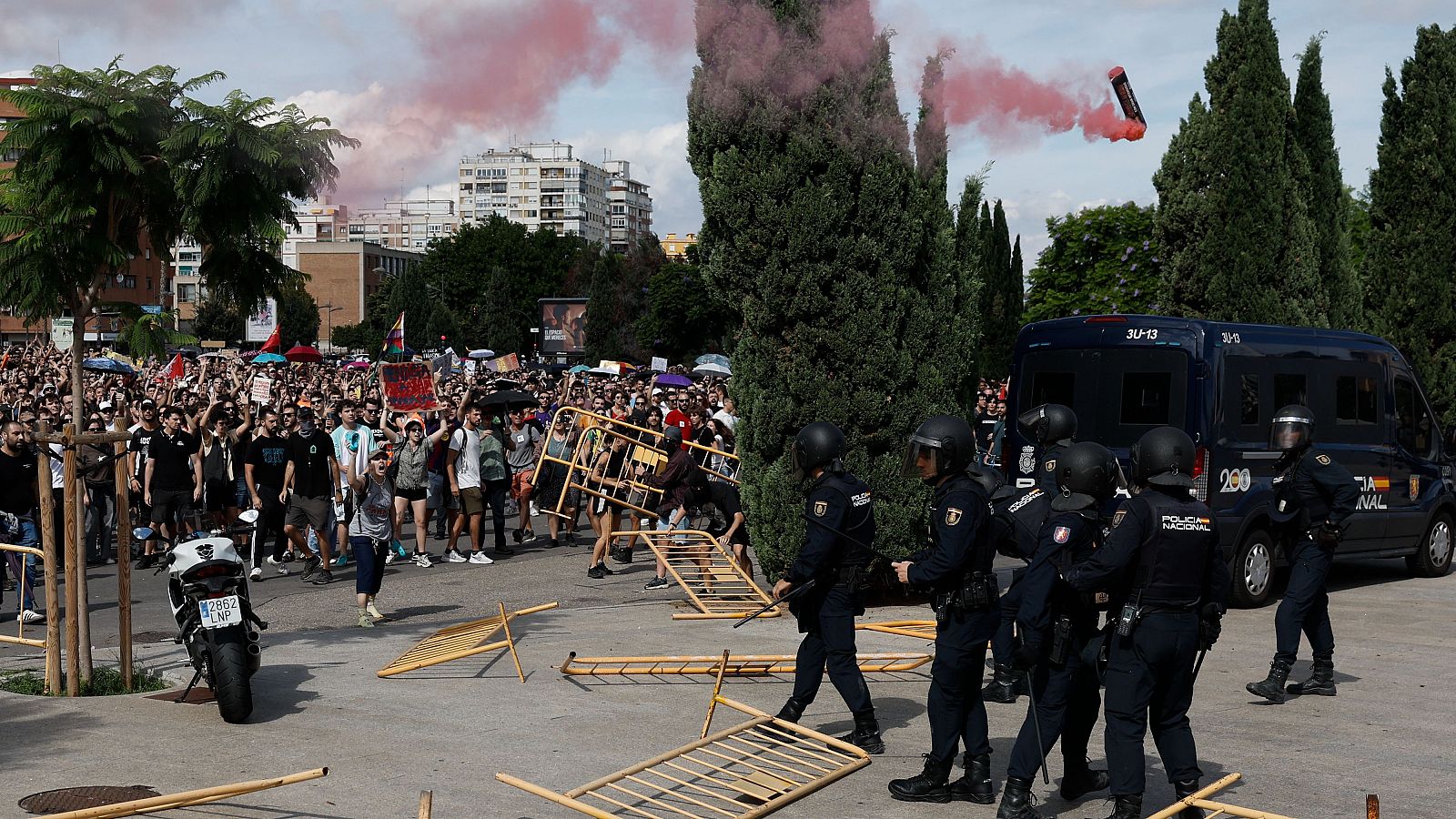 Agentes antidisturbios y un furgón policial junto a vallas caídas en una manifestación urbana con una bengala humeante en el aire y una multitud al fondo.
