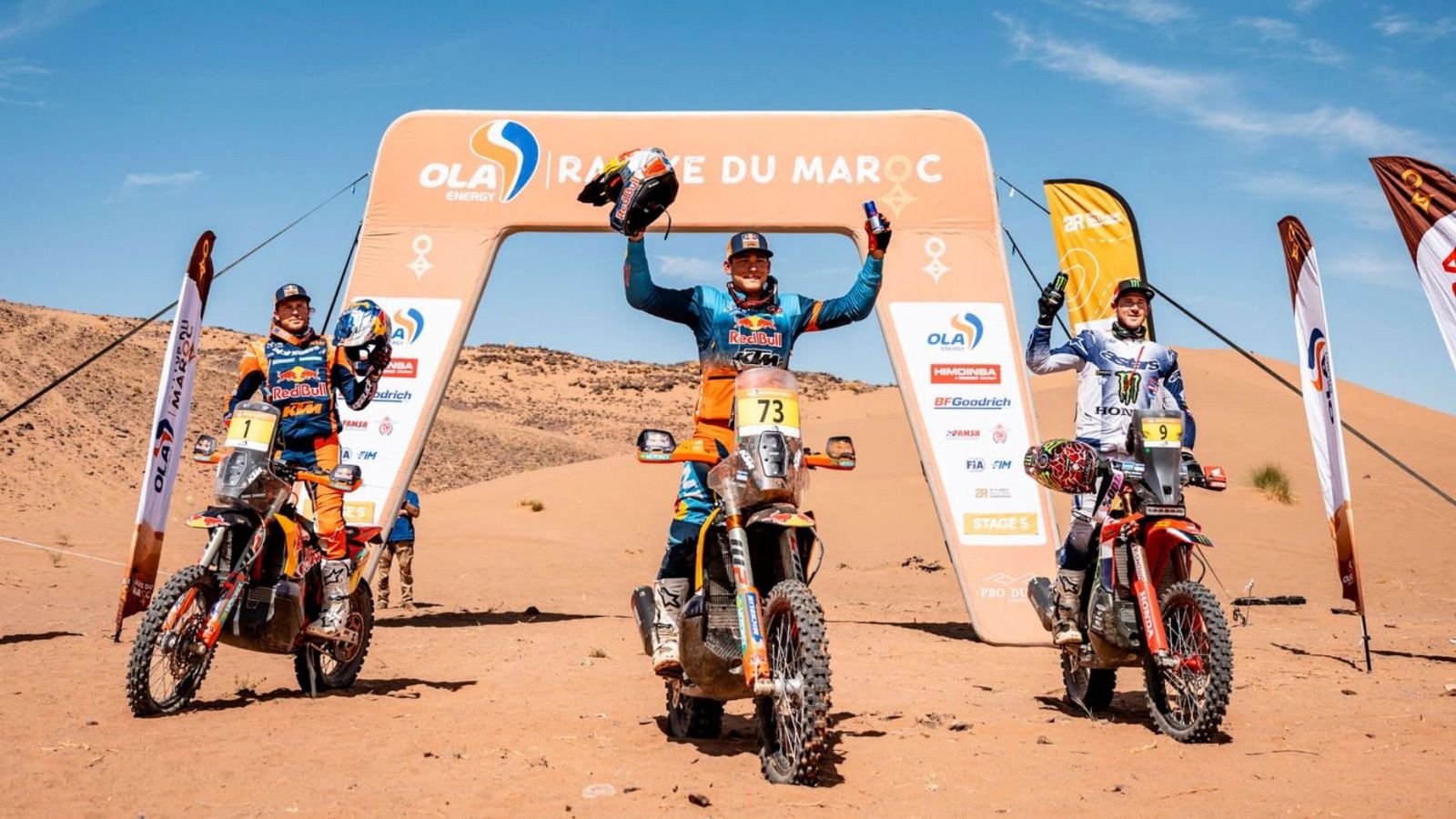 Tres pilotos de rally celebran sobre sus motos en un desierto de arena bajo un arco inflable publicitario, con cielo azul despejado.