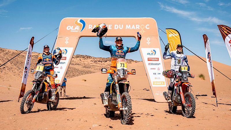 Tres pilotos de rally celebran sobre sus motos en un desierto de arena bajo un arco inflable publicitario, con cielo azul despejado.