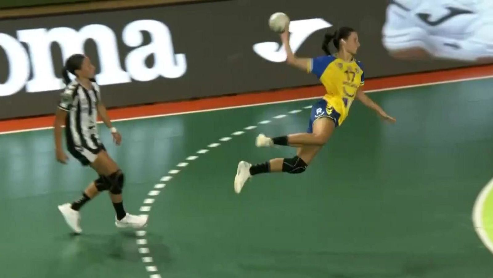 Jugadora de balonmano con el número 17 y camiseta amarilla salta en el aire para lanzar a portería en una pista cubierta con suelo verde y líneas blancas.