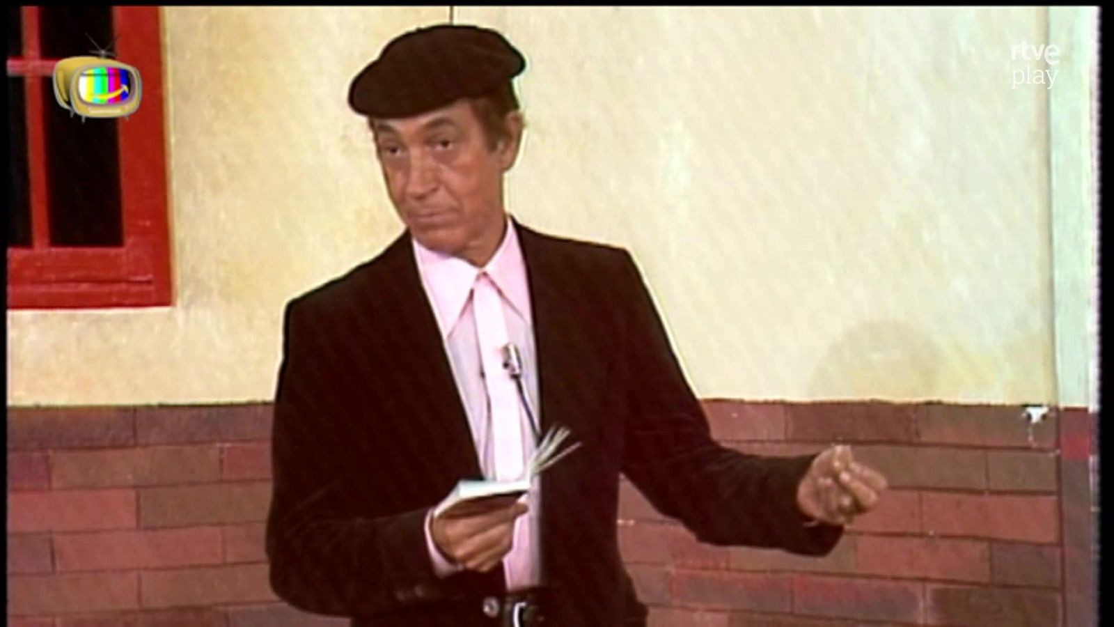 Cómico en un plató televisivo con boina negra, chaqueta oscura y camisa rosa, gesticulando mientras sostiene una libreta ante un fondo de ladrillo y pared beige.