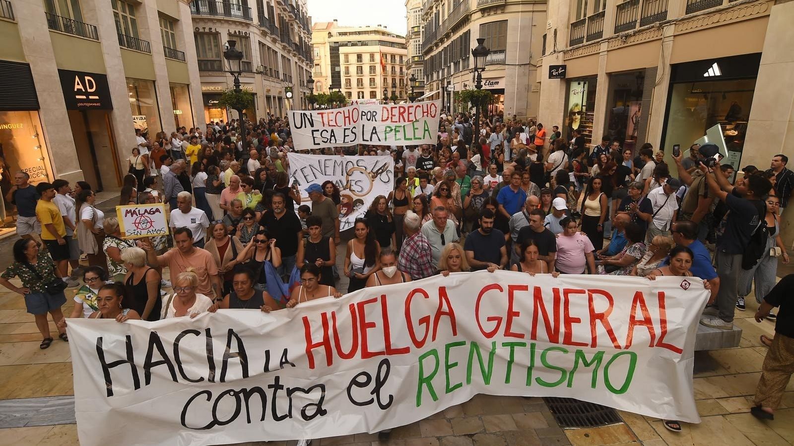 Manifestación ciudadana por el derecho a la vivienda recorriendo una calle peatonal flanqueada por edificios y tiendas, con personas sosteniendo pancartas reivindicativas.