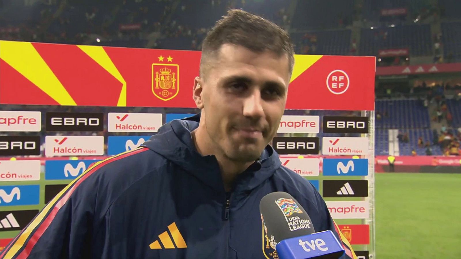 El jugador Rodrigo Hernández concede una entrevista a pie de campo con la equipación oficial ante un panel publicitario y las gradas de un estadio.