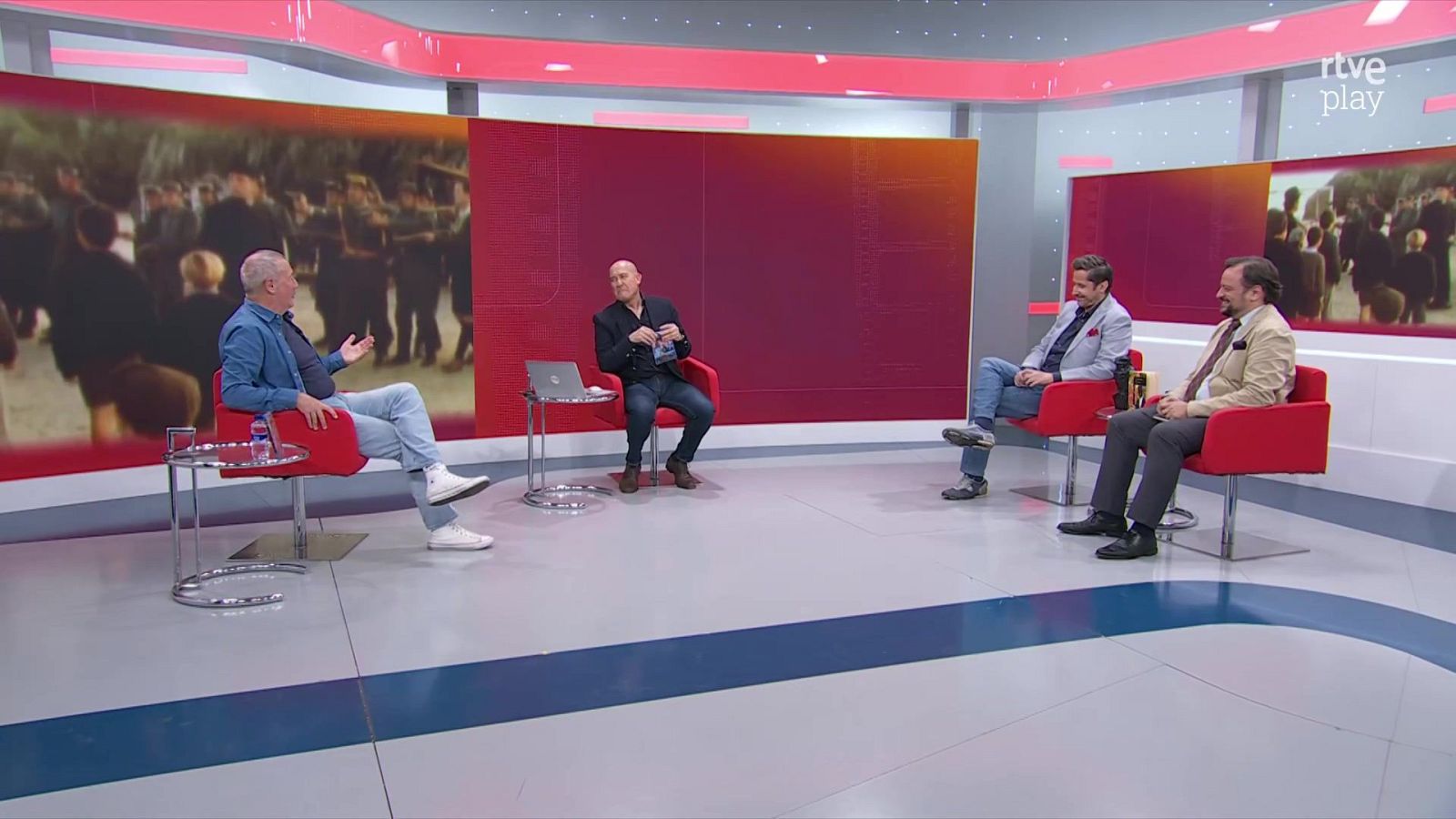 Cuatro hombres participan en una tertulia en un plató de televisión, sentados en sillones rojos sobre un suelo gris y azul, con paneles de fondo que muestran escenas cinematográficas.