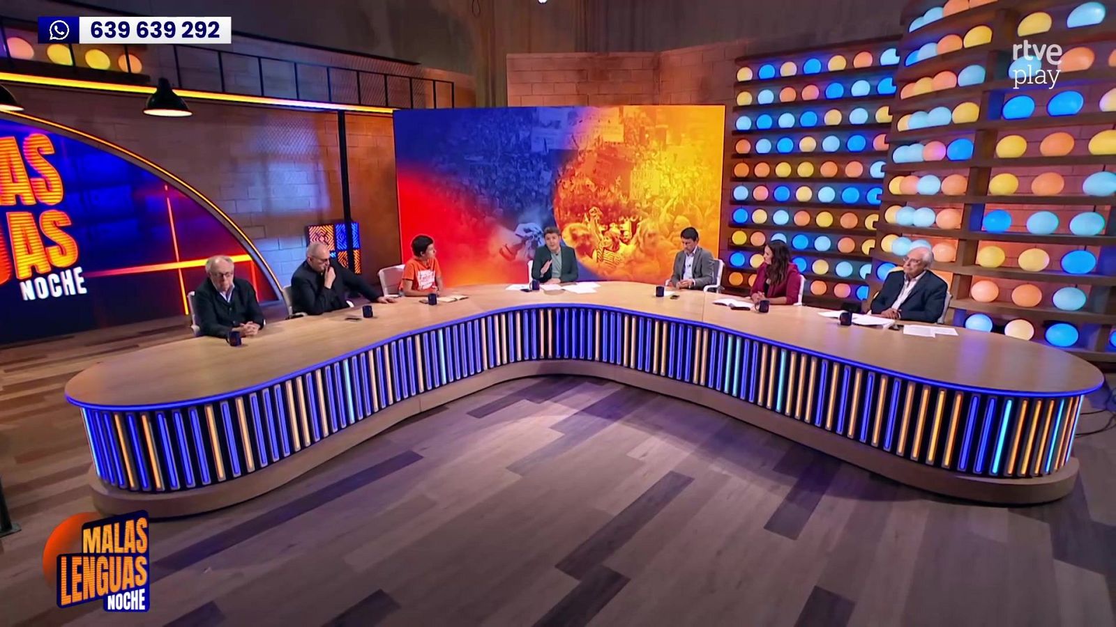Plató de un programa de debate con una mesa semicircular, un moderador, tertulianos, pantallas y elementos gráficos del espacio televisivo.
