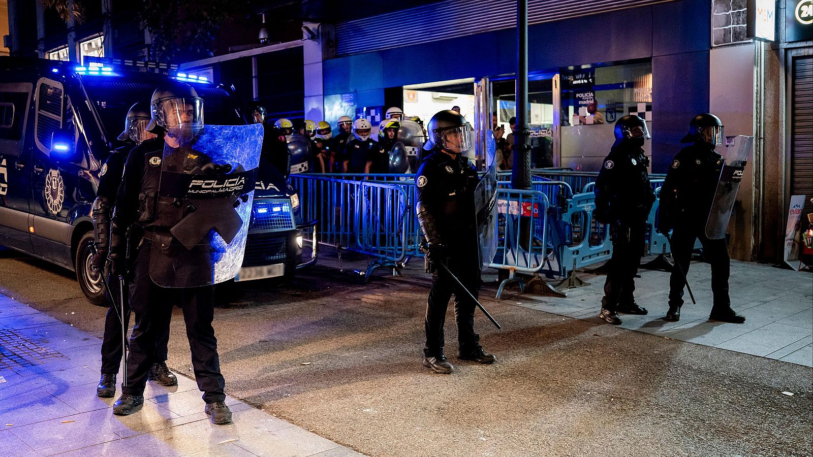 Despliegue nocturno de agentes antidisturbios de la policía con escudos y cascos tras vallas de contención, iluminados por luces de emergencia azules.