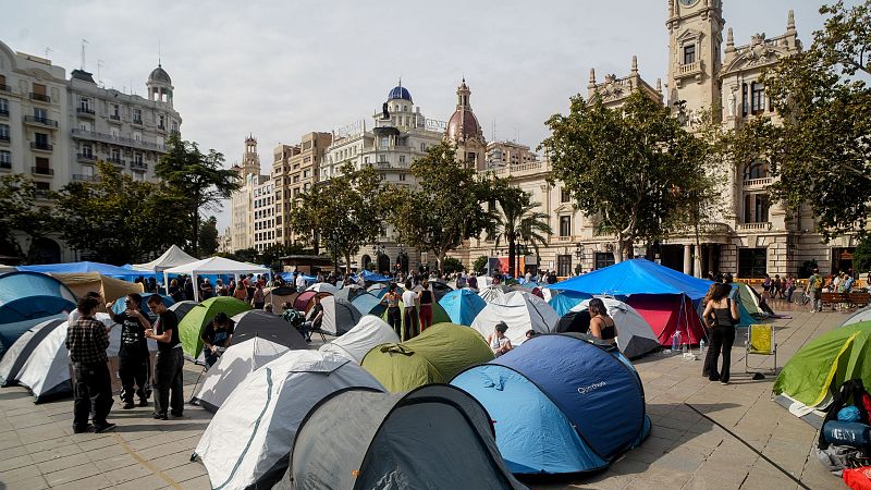 Vista general de una acampada multitudinaria con tiendas de campaña en una plaza urbana junto al edificio del Ayuntamiento.