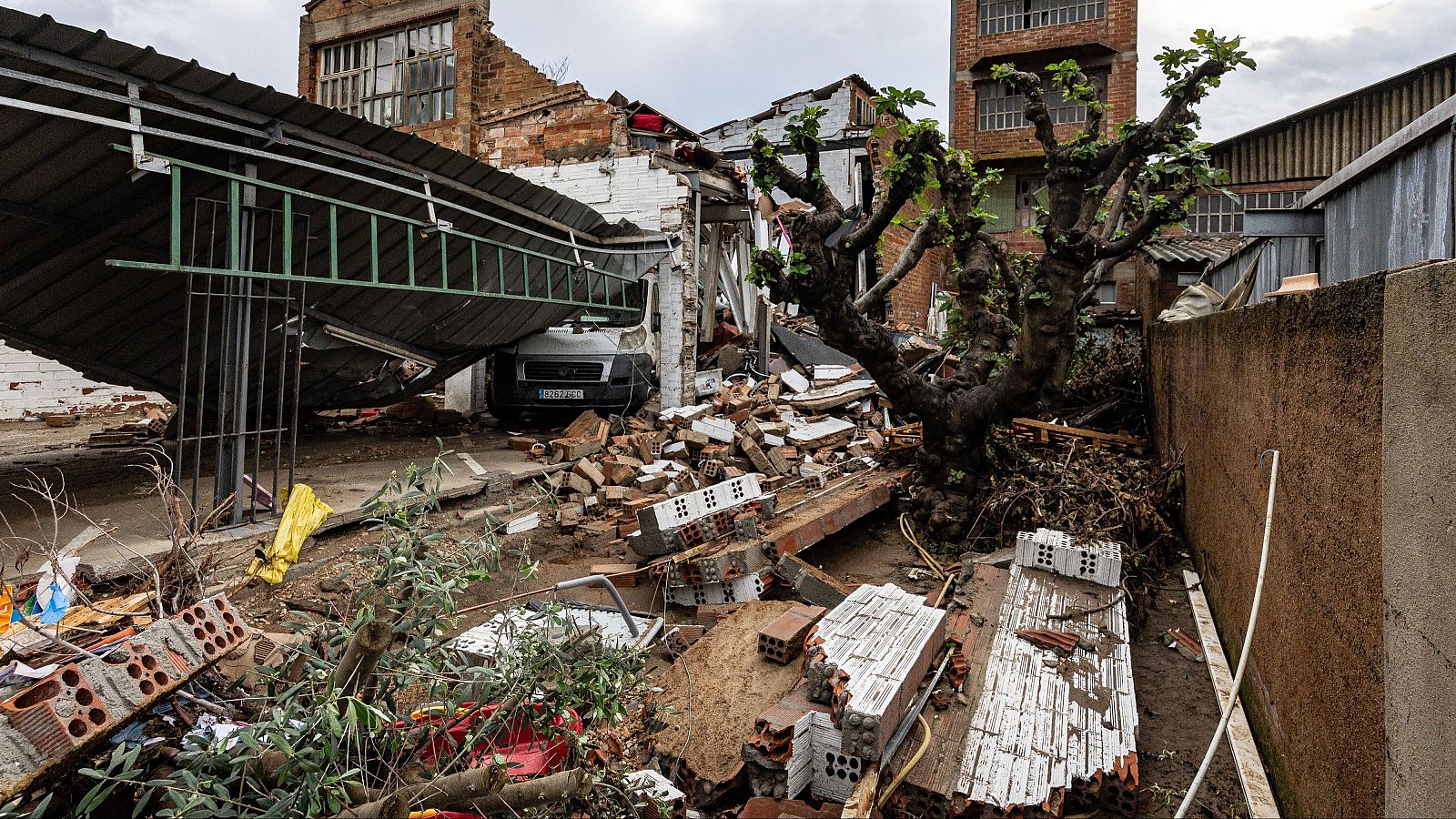 Escenario de destrucción urbana tras un temporal con una marquesina metálica caída, escombros, ladrillos y fango en el suelo junto a un árbol en pie y edificios industriales dañados al fondo.