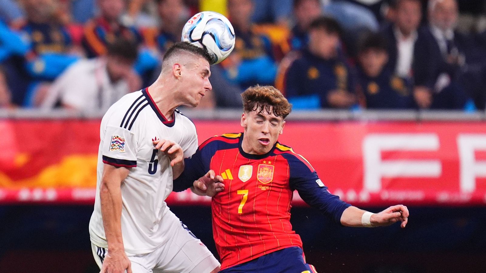 Dos futbolistas de España y la República Checa disputan un balón de cabeza en pleno salto durante un partido oficial, con las gradas del estadio al fondo.