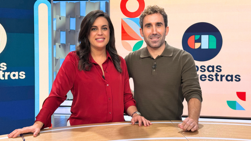 Dos presentadores de televisión están de pie tras una mesa en un plató con pantallas y escenografía geométrica.
