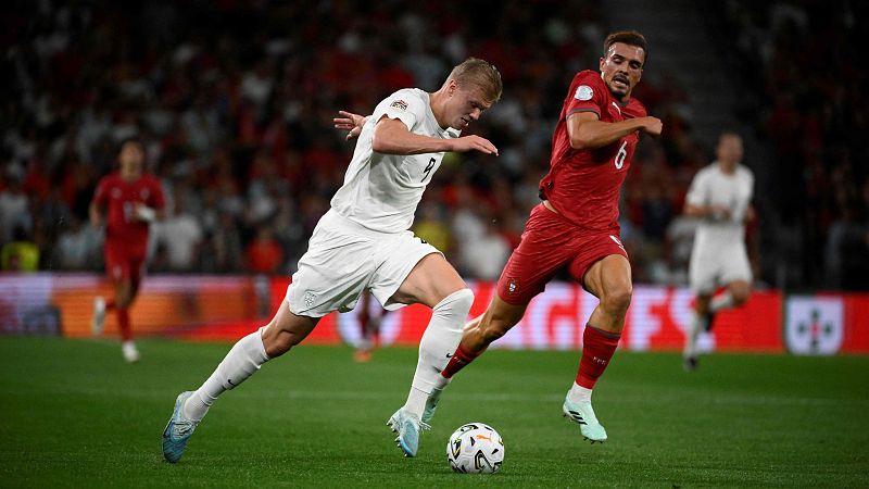 El delantero noruego Erling Haaland con la equipación blanca y el dorsal 9 conduce el balón intensamente, perseguido por un jugador de la selección de Austria sobre el césped del estadio.