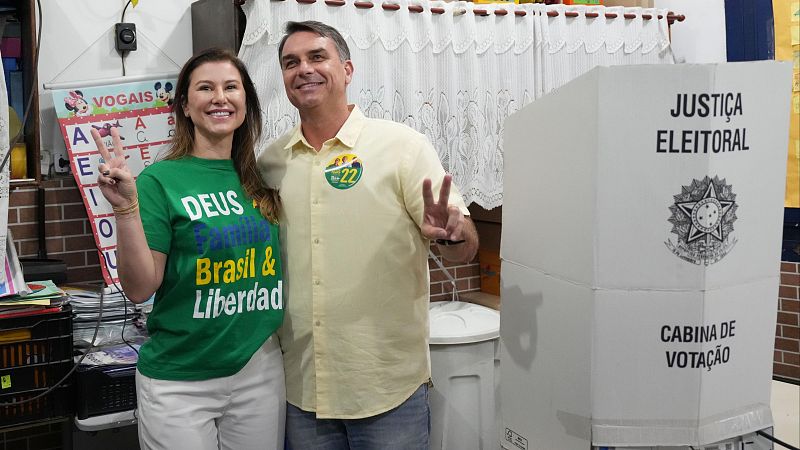 Flávio Bolsonaro y su esposa Fernanda posan sonrientes y haciendo la señal de la victoria junto a una cabina de votación en un colegio electoral.