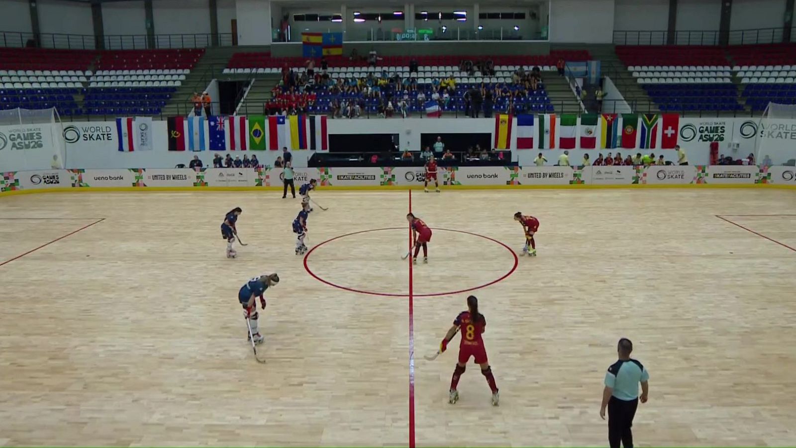 Partido de hockey sobre patines femenino con jugadoras rojas y azules en una pista de parqué y gradas decoradas con banderas internacionales.
