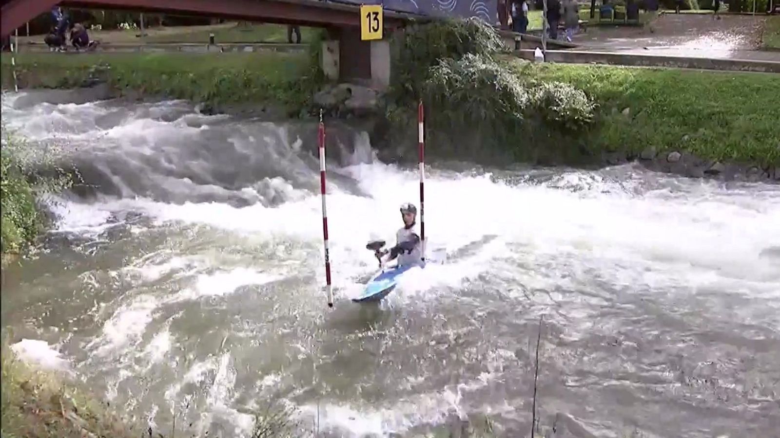 Un palista en kayak navega por aguas bravas entre balizas rojiblancas y bajo un puente con el número 13, observado por espectadores.