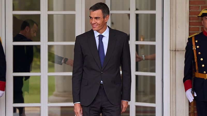 Pedro Sánchez sale al exterior a través de una puerta acristalada vistiendo traje formal y corbata azul marino.