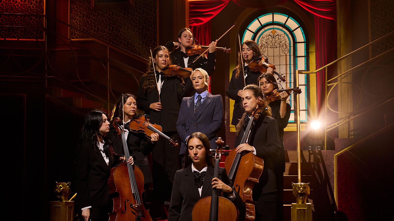 Henar Álvarez posa seria en la escalinata del plató de 'Al cielo con ella', acompañada por una orquesta femenina de instrumentos de cuerda con elegante escenografía clásica.