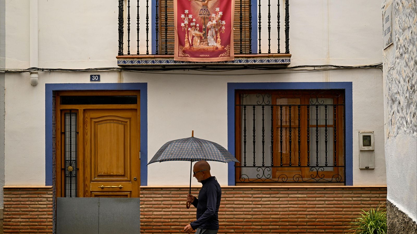 Fachada de casa tradicional en día de lluvia con un hombre caminando con paraguas y un estandarte religioso en el balcón.